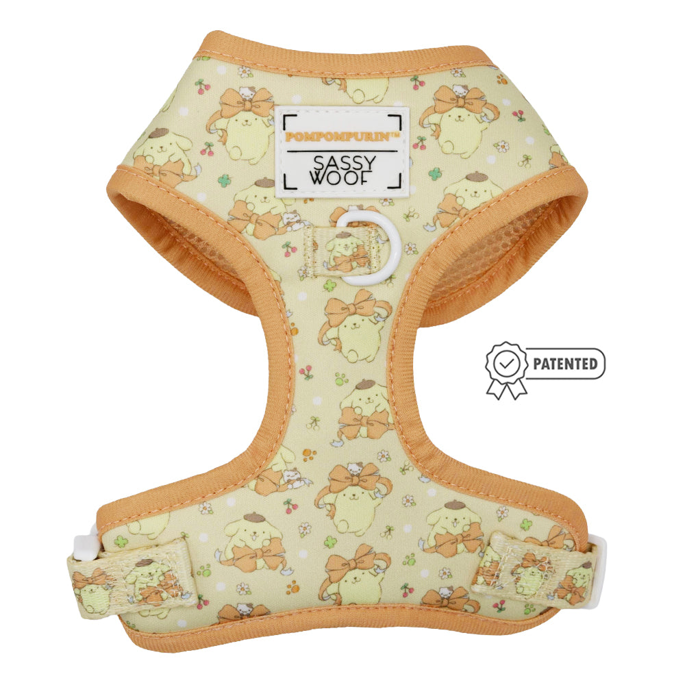 The Dog Adjustable Harness - Pompompurin™ by Sassy Woof features a beige all-over Pompompurin print with flowers and bows, orange trim, white clasp, and branded labels reading “Pompompurin Sassy Woof” and “PATENTED.”.