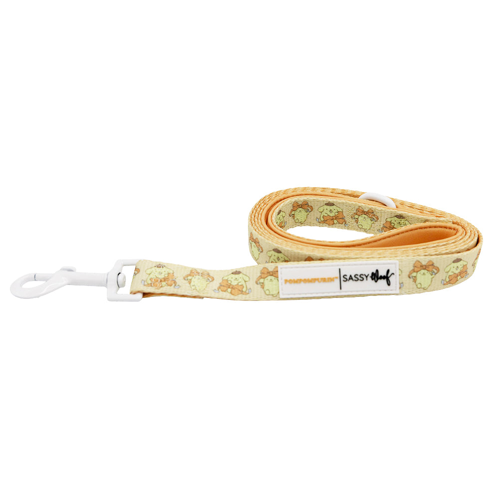 The Dog Leash - Pompompurin™ by Sassy Woof is yellow with cartoon dog faces, a white metal clip, a "POMPOMPURIN SASSY DOG" label, and features a padded neoprene handle for comfort. The coiled leash displays its playful design.