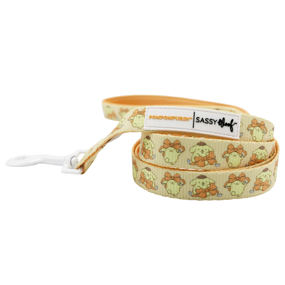 The Sassy Woof Dog Leash - Pompompurin™ is a yellow leash with a cute Pompompurin cartoon holding a croissant, white clasp, branded tags, and padded neoprene handle—pair it with an adjustable harness for stylish walks.
