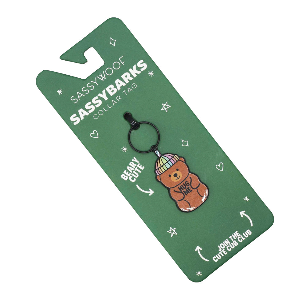 A green card labeled Sassy Woof Dog Collar Tag - Hug Me Bear displays a pastel-beanie bear tag keychain with “Hug Me” on its belly, “Berry Cute,” and “Join the Cute Cub Club”—the sweetest dog tag accessory for your pup!.