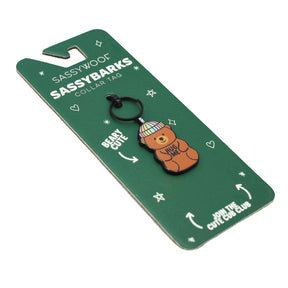 A green Sassy Woof Collar Tag - Hug Me Bear package displays a bear charm with a pastel beanie and "HUG ME." The packaging features stars and phrases like "BEARY CUTE" and "JOIN THE CUTE CUB CLUB.