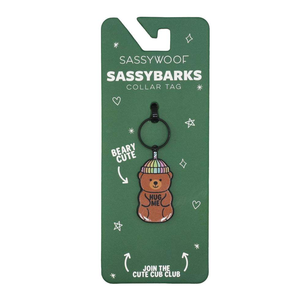 The Sassy Woof Dog Collar Tag - Hug Me Bear features a pastel bear wearing a striped hat with "Hug Me" on its belly, displayed on playful green Sassy Woof packaging with fun text and star doodles.