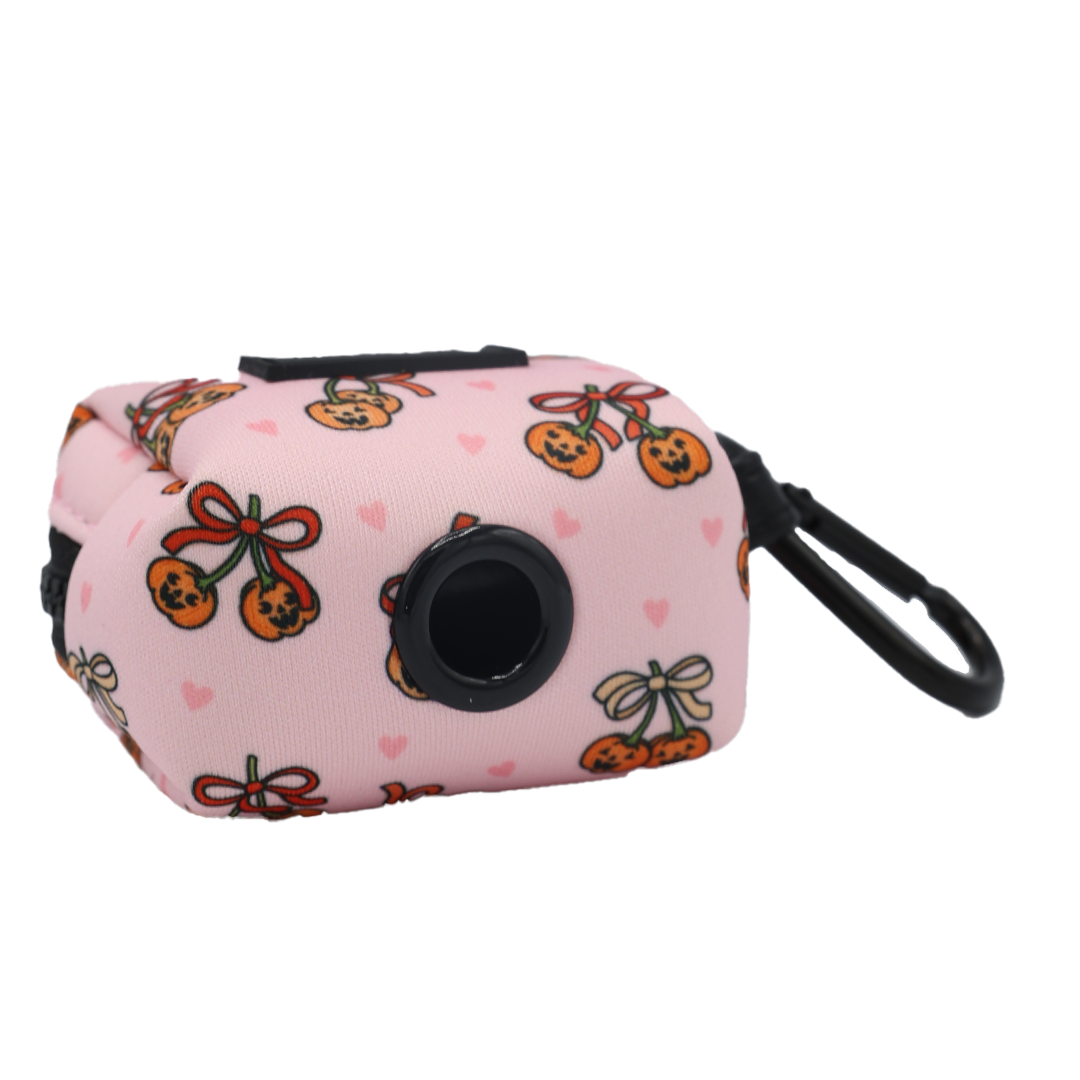 Introducing the Dog Waste Bag Holder - Sassy Lil Pumpkins by Sassy Woof—a pink accessory from the Sassy Lil Pumpkins collection, decorated with orange cherries, bow designs, and small pink hearts. It features a convenient black dispenser opening and a sturdy black clip for easy attachment, making it the perfect fun pumpkin-themed dog accessory.