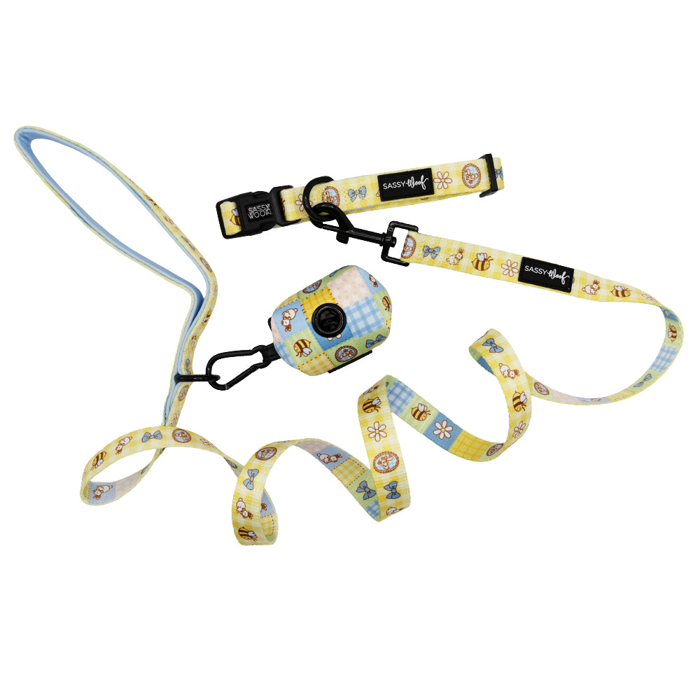 The Sassy Woof Dog Three Piece Bundle - Patch Perfect features a playful bee and flower picnic pattern in yellow and blue, with black clasps and signature tags—perfect for stylish pups! Includes a matching leash, collar, and waste bag holder.