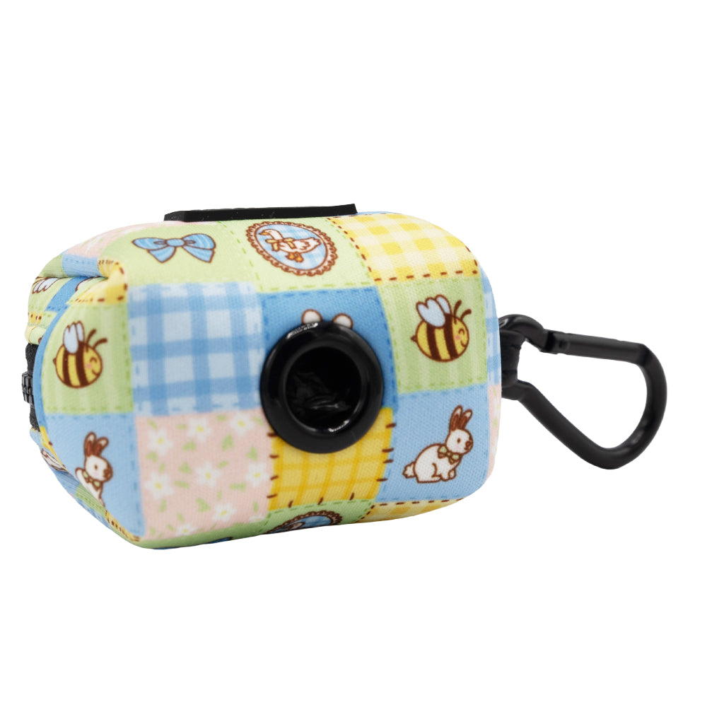 The Sassy Woof Dog Four Piece Bundle - Patch Perfect includes a small, rectangular, pastel patchwork bag dispenser with bees, bunnies, and bows, a black clip, and a front black plastic opening for dispensing bags.