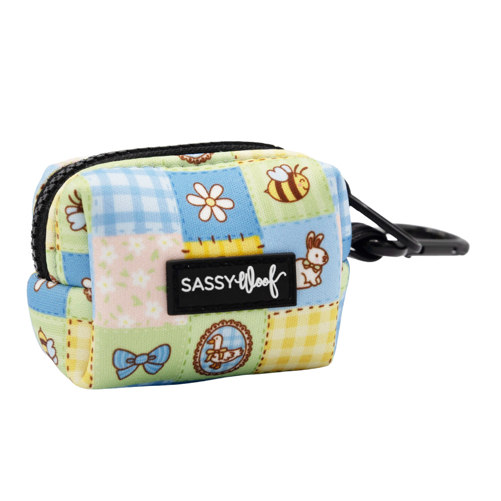The Sassy Woof Dog Waste Bag Holder - Patch Perfect is a small, square neoprene holder with zipper and clip, featuring pastel quilted patterns of flowers, bees, bunnies, bows, a basket, and finished with a front Sassy Woof label.