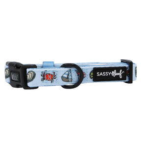 The Sassy Woof Coastal Cuties dog collar features light blue fabric with nautical designs (sailboat, seashell, crab), black hardware, and a small brand logo patch—ideal for fans of coastal-themed pet accessories.
