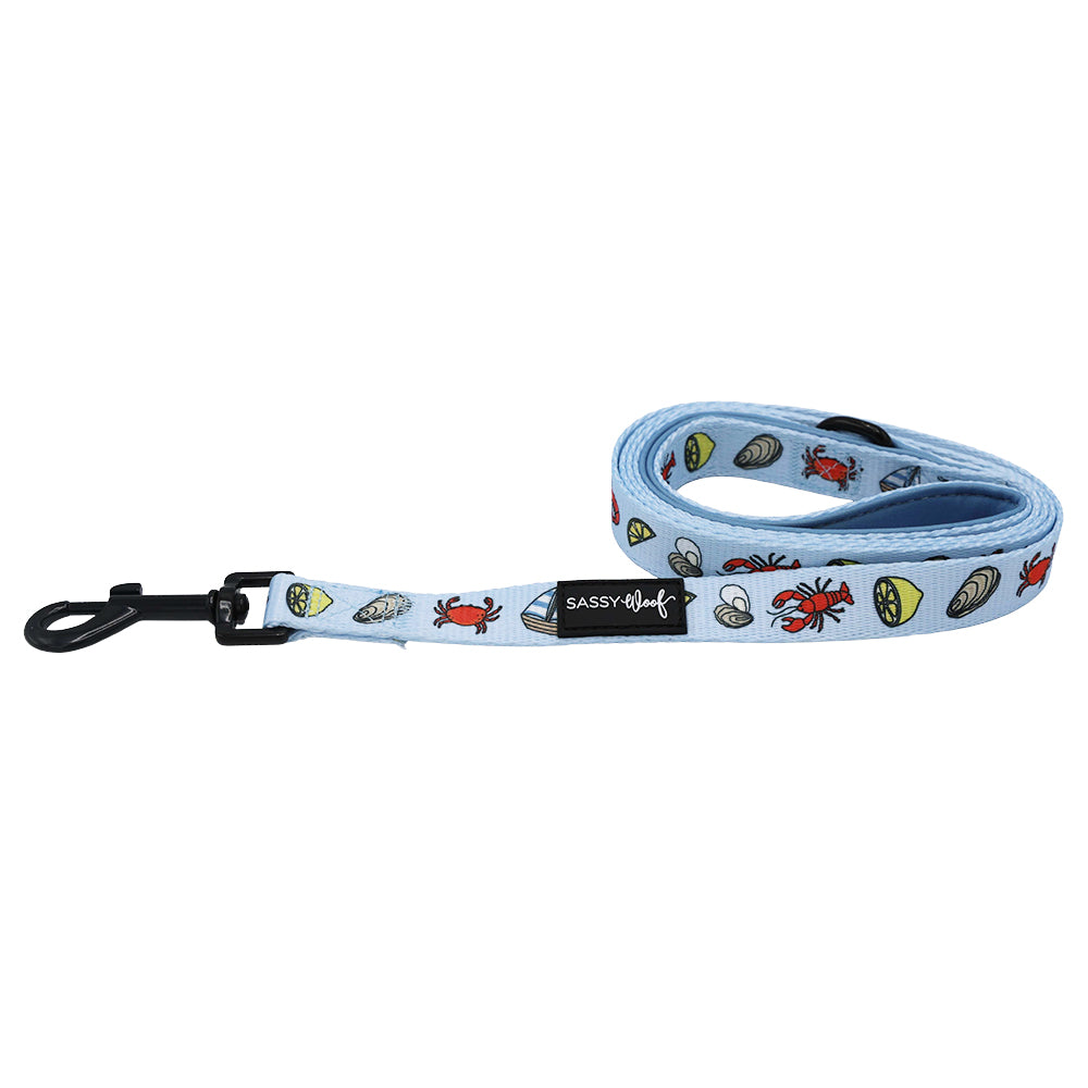 The Sassy Woof Dog Leash - Coastal Cuties is light blue with colorful seafood prints and a black clip. It matches the adjustable harness and features a signature Sassy Woof label for extra charm.