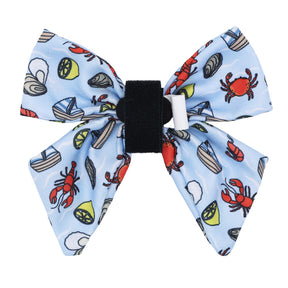The Sassy Woof Dog Sailor Bow - Coastal Cuties is a blue bowtie with a black center band, featuring red crabs, lemon slices, oysters, sailboats, and seashells in a playful seaside pattern.