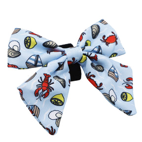 The Sassy Woof Dog Sailor Bow - Coastal Cuties is a light blue bow featuring red lobsters, crabs, lemon slices, clams, and oyster shells on a black band.