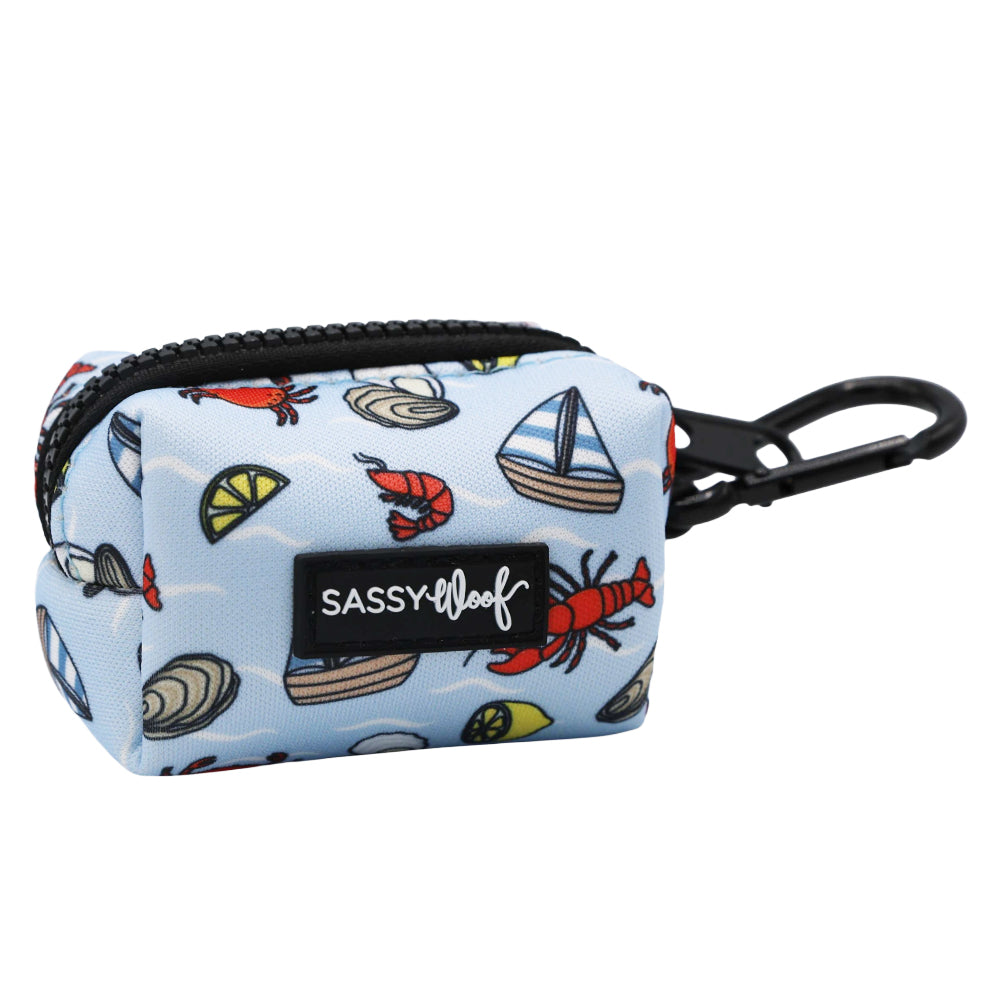 The Sassy Woof Dog Waste Bag Holder - Coastal Cuties is a small, light blue neoprene holder with a black clip, featuring red lobsters, clams, lemon slices, sailboats, and a black Sassy Woof label—ideal for coastal adventures!.