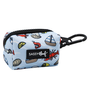 The Sassy Woof Dog Waste Bag Holder - Coastal Cuties is a small, light blue neoprene holder with a black clip, featuring red lobsters, clams, lemon slices, sailboats, and a black Sassy Woof label—ideal for coastal adventures!.