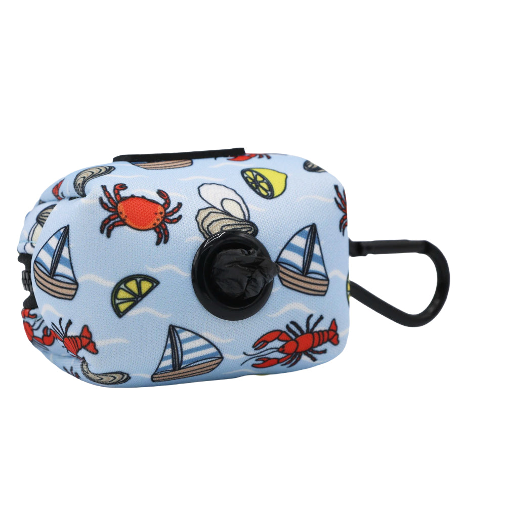 The Dog Waste Bag Holder - Coastal Cuties by Sassy Woof is a small, rectangular blue neoprene holder with cheerful nautical prints. It includes a black dispenser and clip, making it perfect for stylish pet owners on the go.