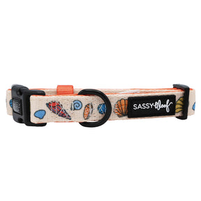 The Sassy Woof Dog Collar - Shell Yeah features a beige, ocean-inspired design with seashells and starfish, a black buckle, adjustable strap, metal ring, and a black SASSY WOOF patch in white lettering.