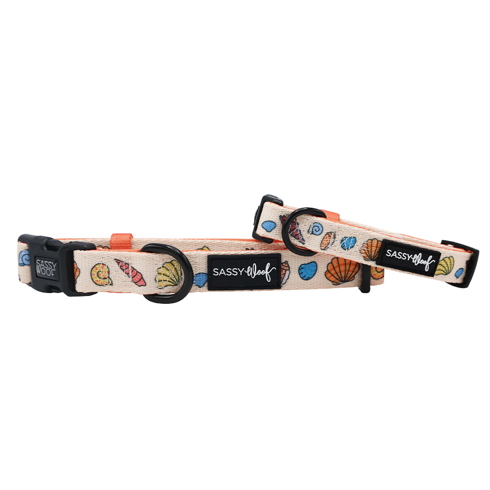 Two adjustable Sassy Woof "Shell Yeah" dog collars feature colorful seashell designs on a light background, black buckles, and neoprene material. Each collar includes a black Sassy Woof logo tag for added style and comfort.