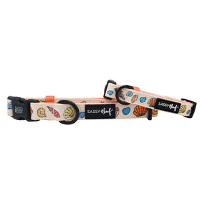 Two adjustable Sassy Woof "Shell Yeah" dog collars feature colorful seashell designs on a light background, black buckles, and neoprene material. Each collar includes a black Sassy Woof logo tag for added style and comfort.