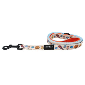 The Sassy Woof "Shell Yeah" dog leash features a beige seashell design with blue trim, orange underside, black metal clasp, and a Sassy Woof label—ideal for beach-loving pups and their owners.