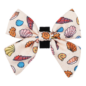 The Sassy Woof Dog Sailor Bow - Shell Yeah features a large fabric bow with pink, blue, orange, and yellow shells and starfish on a light background and includes a black center clip for easy attachment to your dog's collar.