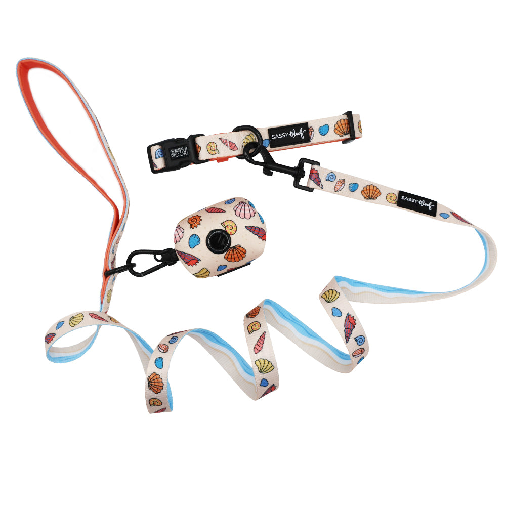 The Sassy Woof Dog Three Piece Bundle - Shell Yeah features a leash, collar, and waste bag holder with colorful seashells on white, accented in light blue and orange—perfect for pups who love seaside style!.