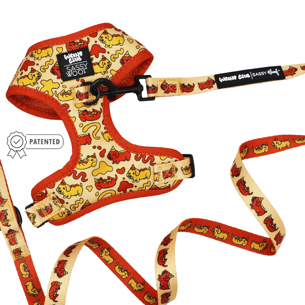 The Sassy Woof Dog Two Piece Bundle - Hot Dawg includes a yellow and orange adjustable harness and matching leash with playful dog patterns, plus a patented badge to the left of the harness.