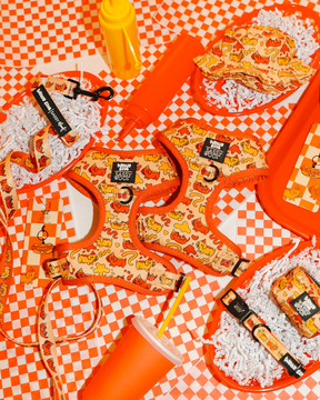 Sassy Woof’s Dog Four Piece Bundle - Hot Dawg, featuring a dachshund harness with orange and yellow cartoon prints, is displayed on a red-and-white checkered surface with playful diner-themed decor.