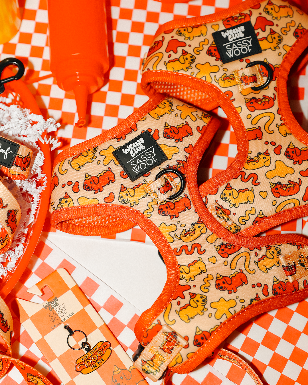 Two Sassy Woof "Hot Dawg" adjustable dog harnesses with playful orange and yellow designs are displayed on a checkered surface, accompanied by a ketchup bottle, gift tag, and shredded paper for a fun, vibrant display.