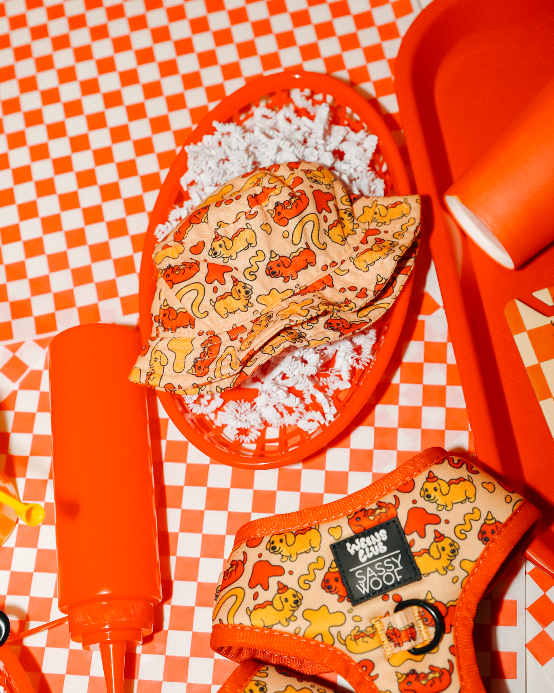 The Sassy Woof Dog Bucket Hat - Hot Dawg, featuring a vibrant food-themed print, rests on shredded paper in a red tray, alongside an adjustable harness, ketchup bottle, and red cup atop a red-and-white checkered tablecloth.
