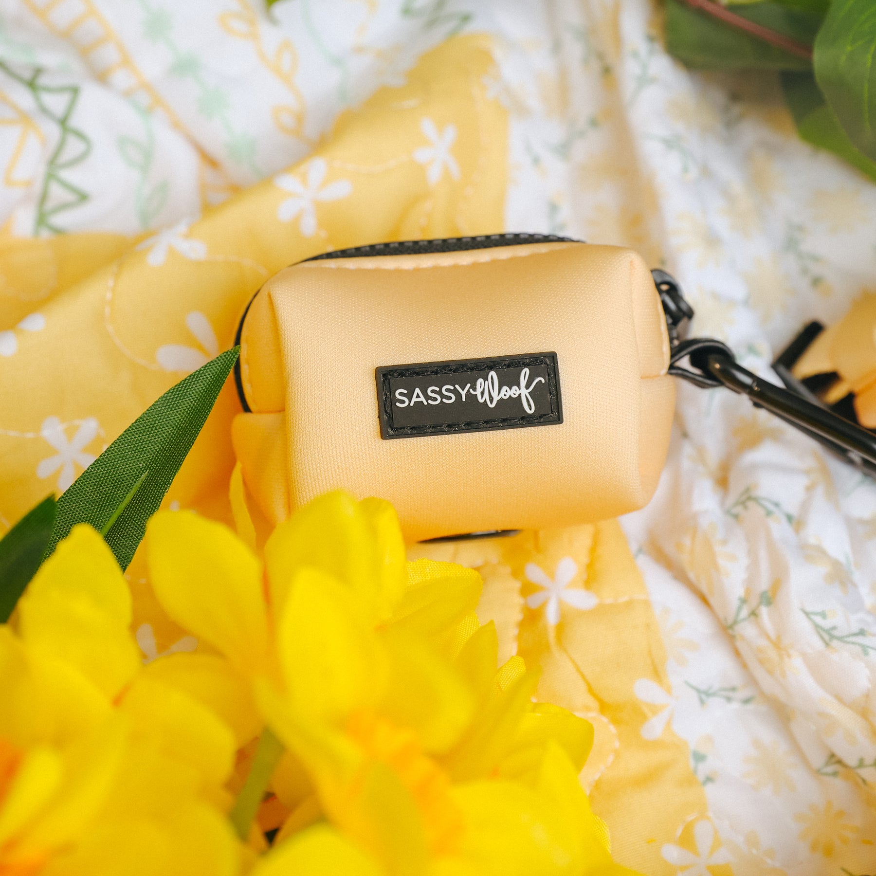 The Sassy Woof Dog Three Piece Bundle - Daffodil sits on yellow floral fabric, surrounded by bright yellow flowers and green leaves.
