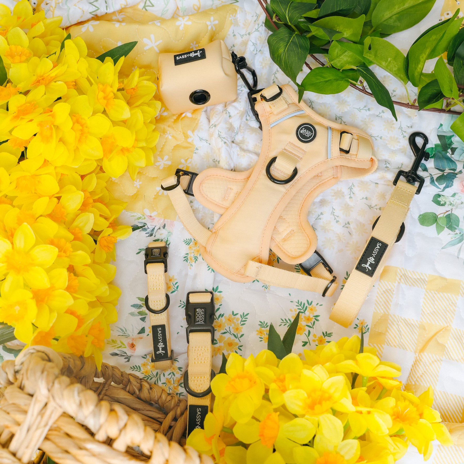 The Sassy Woof Dog Four Piece Bundle - Daffodil includes a pastel yellow harness, leash, collar, and waste bag holder, beautifully showcased on a floral picnic blanket with bright yellow flowers and a woven basket.