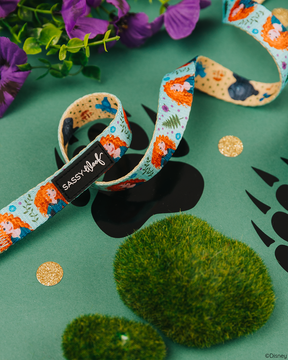 A decorative ribbon with cartoon characters sits on a green surface with black paw prints, moss, glitter dots, and purple flowers—perfect to pair with the Sassy Woof Dog Leash - Disney's Merida. The ribbon displays a Sassy Woof label.
