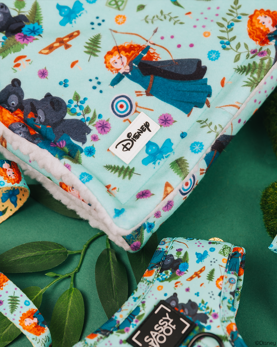 The Sassy Woof Pet Blanket - Disney's Merida features Princess Merida, bears, arrows, targets, and forest elements. Cozy sherpa fabric with Disney and Sassy Woof labels; accented by green leaves and moss for dogs.