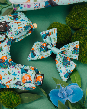 A light blue Sassy Woof Dog Sailor Bow inspired by Disney’s Merida, displayed with green moss, leaves, and a blue orchid on a green background.