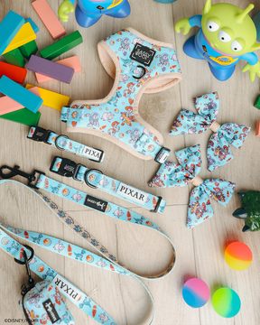 The Sassy Woof Dog Four Piece Bundle – Disney and Pixar's Toy Story is shown on a wooden floor with colorful blocks and playful Toy Story alien figures. The set includes an adjustable harness, leash, collar, and bows featuring beloved characters.