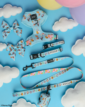 The Sassy Woof Dog Four Piece Bundle – Disney and Pixar's Up includes an adjustable harness, leash, collar, and waste bag holder in pastel blue, shown on a sky-blue background with clouds and pastel balloons.