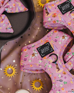 The Sassy Woof Dog Two Piece Bundle – Disney’s Rapunzel features a pink pet harness with castles, Pascal, purple flowers, and gold sun motifs, plus a matching bow in a cast iron pan and sheer fabric accents.