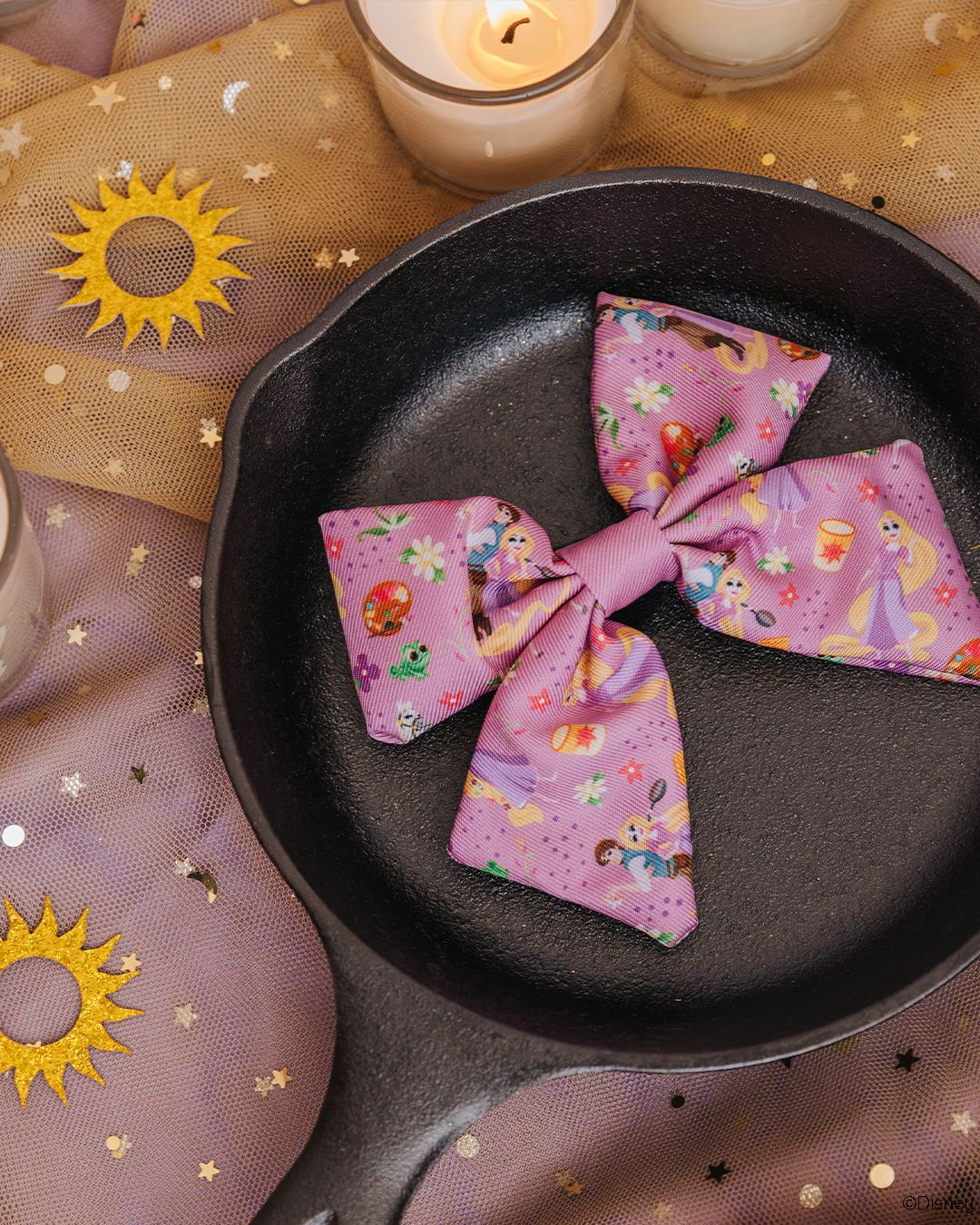 A Sassy Woof Dog Sailor Bow – Disney’s Rapunzel sits in a black cast iron pan, surrounded by glowing candles and metallic sun and star decorations atop purple mesh fabric.