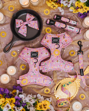 A flat lay of the Sassy Woof Harness Three Piece Bundle - Disney's Rapunzel, with pink harnesses, leash, and collar, styled with candles, pansies, faux jewels, a frying pan, and a gold star-patterned cloth.