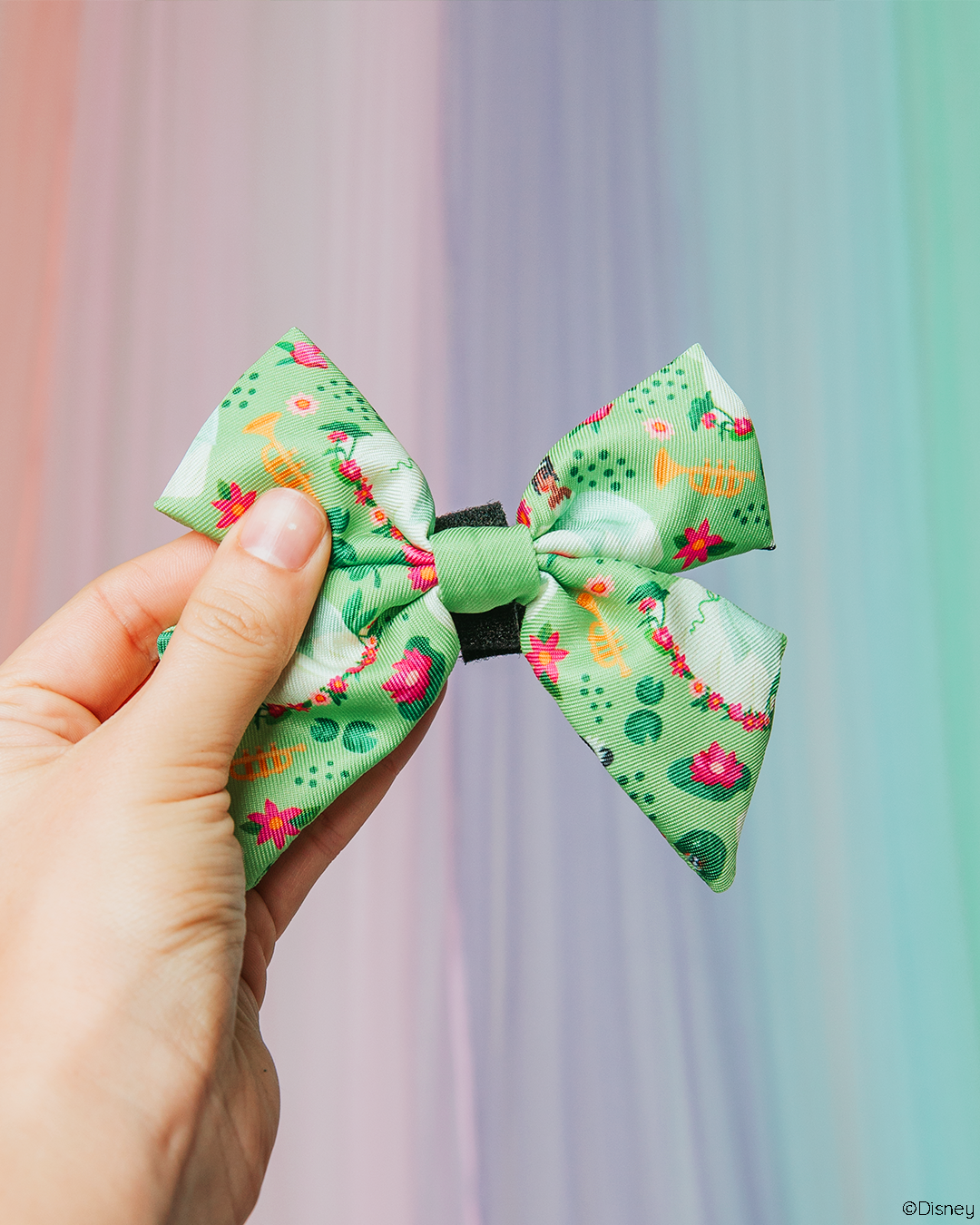 A hand holds the Sassy Woof Dog Sailor Bow - Disney's Tiana, a green hair accessory with colorful flowers and animals, styled like a Princess and the Frog dog harness, against a pastel rainbow gradient background.
