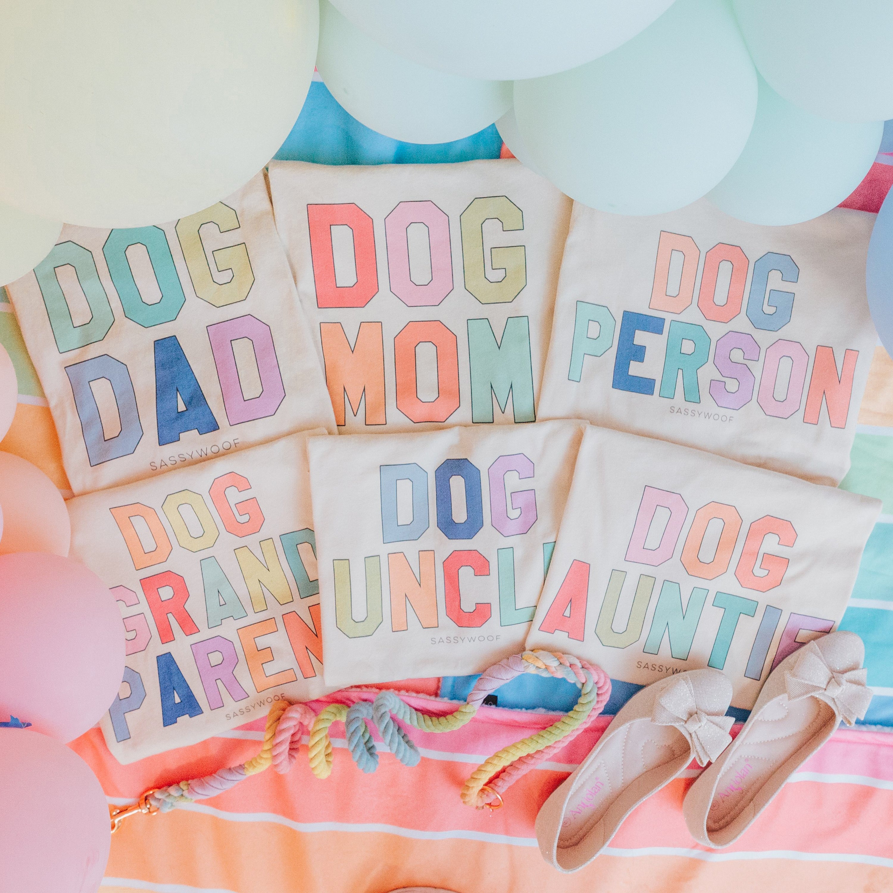 Five pastel tote bags with phrases: Dog Dad, Dog Mom, Dog Person, Dog Grandparent, and Dog Auntie. Surrounding them are balloons, a pastel rope toy, and a pair of beige shoes on a striped blanket.