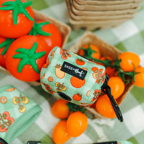The Sassy Woof Dog Three Piece Bundle - Feeling Vine features a green, plant-inspired bag holder with a tomato pattern and label, set among orange tomatoes on a green and white checkered tablecloth—perfect for pups Feeling Vine.