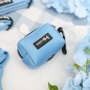 A small, rectangular blue Sassy Woof pouch rests on a white surface with blue and white checkered fabric and light blue flowers, perfectly complementing the Dog Three Piece Bundle - Hydrangea from Sassy Woof.