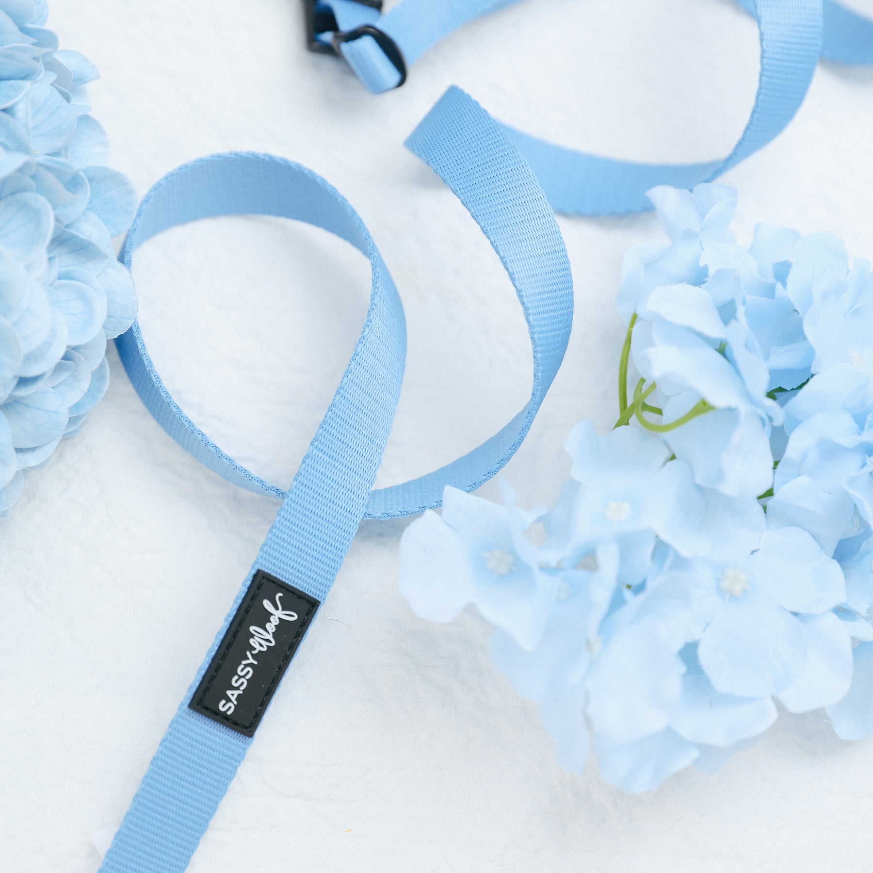 The Sassy Woof Dog Leash - Hydrangea, featuring a black brand tag, is displayed on a white surface beside light blue hydrangea flower clusters.