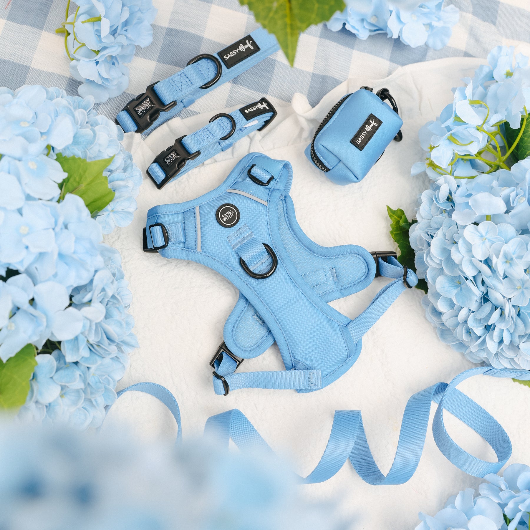 The Sassy Woof Dog Four Piece Bundle - Hydrangea, featuring a light blue harness, leash, and waste bag holder, is displayed on a white blanket with blue ribbon amid pastel hydrangeas and a blue checkered cloth.