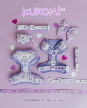 A pastel purple Sassy Woof Dog Collar - Kuromi™ set is shown on a lavender heart background, with an adjustable collar, two harnesses, bow, leash, waste bag holder, and decorative tape with cute heart patterns.