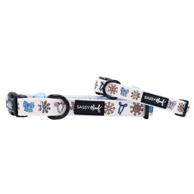 Two white Sassy Woof "Let it Bow" dog collars with black buckles and winter designs—snowflakes, mittens, hats, gifts. Each features a black Sassy Woof tag and pairs perfectly with a blue bow collar for extra seasonal style.
