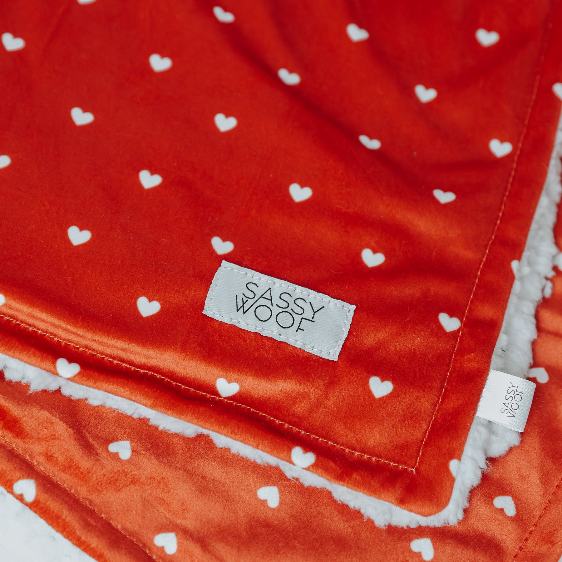 A close-up of the Sassy Woof Pet Blanket - Little Love shows its red velvet fabric with white hearts, a fluffy white edge, and a sewn Sassy Woof label in the corner, all part of the Valentine’s collection.