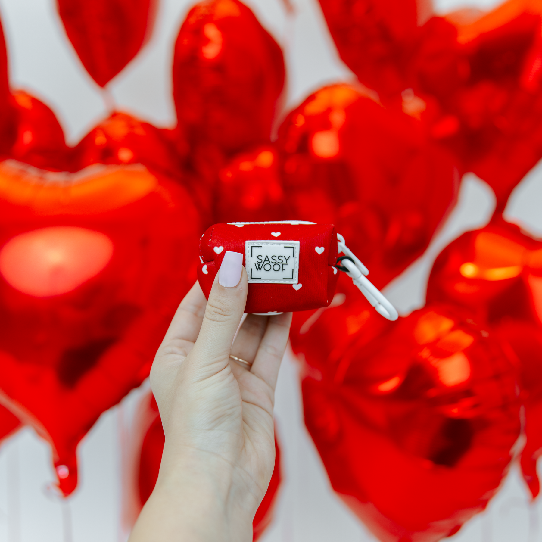 Hand holding a red AirPod case with 'Sassy Wool' branding against a heart-patterned background.