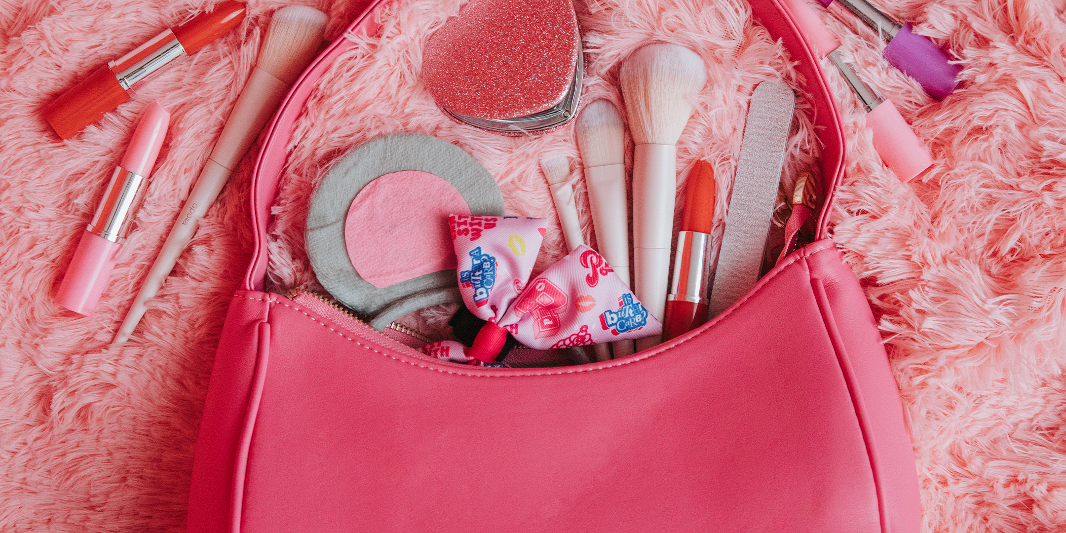 Pink makeup bag with various makeup items on a pink textured surface