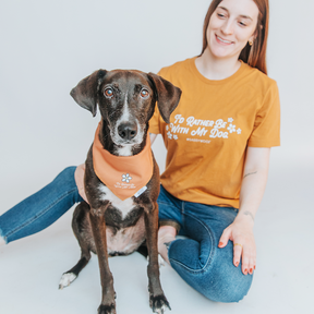 A dog mom wears the Sassy Woof Human Tee - I’d Rather Be With My Dog while sitting beside her black dog in an orange bandana. Both face the camera against a plain white background.