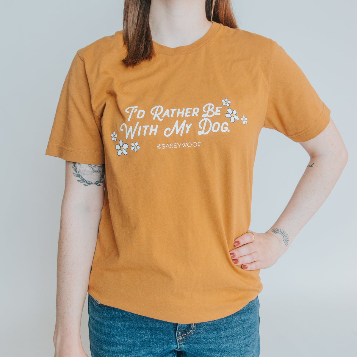 Wearing the Sassy Woof Human Tee - I’d Rather Be With My Dog in orange with white text and paw prints, a tattooed person stands with one hand on their hip.